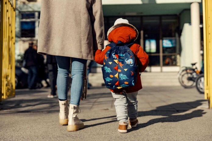 shutterstock_2100589987 Child with dinosaur backpack walking beside adult